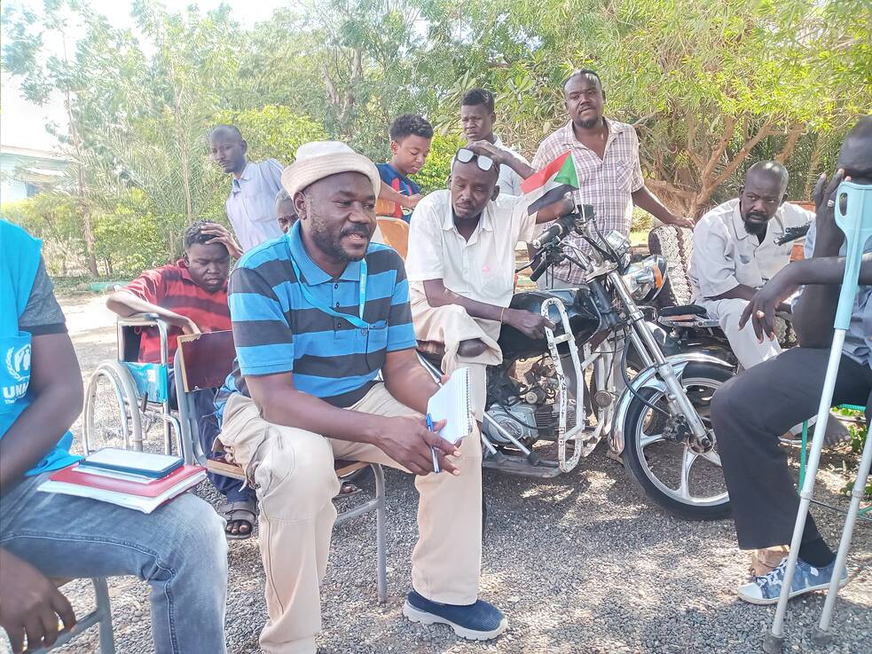 Eine Gruppe von Menschen sitzt teils auf Stühlen, teils in Rollstühlen um einen Mann herum, der einen Schreibblock und Stift in den Händen hält. Hinter ihnen sind Sträucher. 