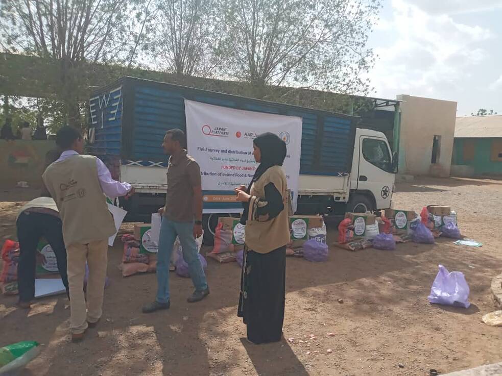 Eine Frau mit einem Notizblock in den Händen, die einen Hidschab trägt, beobachtet zwei Männer, die einen Lastwagen beladen.