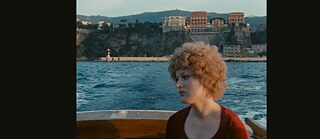 A white woman with reddish-blonde hair in a boat against a backdrop of blue sea and coastal landscape