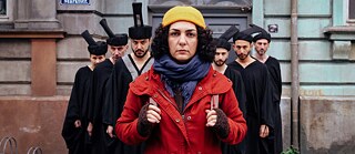 Szene aus dem Film SHAHID: Eine Frau in gelber Mütze und roter Jacke steht vor einer Gruppe schwarz gekleideter Männer.