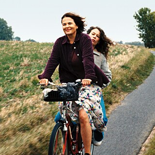 Zwei Frauen in Freizeitkleidung und Röcken fahren mit dem Fahrrad auf einer Landstraße, hinter ihnen Felder und blauer Himmel. 