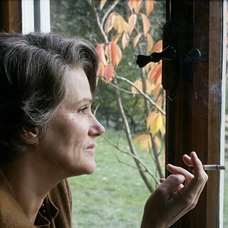 Portrait von Hanna Arendt. Sie sitz im Innenraum an einem Fenster und blickt nach draußen auf herbstlich gefärbte Blätter. In der Hand hält sie eine brennende Zigarette, während vor dem Fenster ein Garten und ein Holzstapel zu sehen sind. Die warme Lichtstimmung erzeugt eine ruhige, nachdenkliche Atmosphäre.