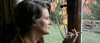Portrait von Hanna Arendt. Sie sitz im Innenraum an einem Fenster und blickt nach draußen auf herbstlich gefärbte Blätter. In der Hand hält sie eine brennende Zigarette, während vor dem Fenster ein Garten und ein Holzstapel zu sehen sind. Die warme Lichtstimmung erzeugt eine ruhige, nachdenkliche Atmosphäre.
