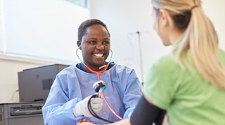Zoleka Hofmann bei der Ausbildung zur Krankenpflegerin. Sie untersucht eine Patientin und misst den Puls. Sie lacht freundlich. 