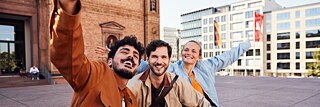 Eine Gruppe von drei Personen posiert fröhlich für ein Selfie vor einem beeindruckenden Gebäude