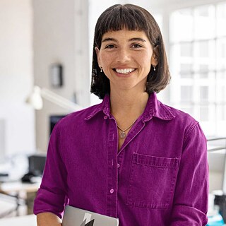 Eine Frau in einem violetten Hemd lehnt sich an einen Schreibtisch in einem Büroraum.