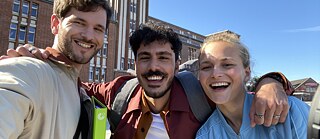 Drei Jugendliche stehen Arm in Arm vor dem Goethe-Institut Hamburg und lachen in die Kamera.