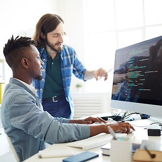 Zwei Personen an einem Schreibtisch mit Computerbildschirm, auf dem Programmiercode angezeigt wird; eine Person sitzt, die andere steht und zeigt auf den Bildschirm. 