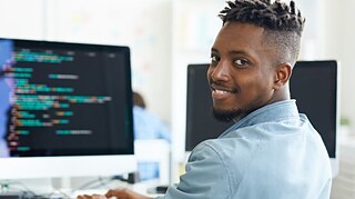 Person mit kurzem, lockigem Haar sitzt an Schreibtisch vor zwei Bildschirmen, auf einem ist Programmiercode sichtbar. 