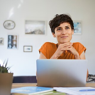 Eine Frau trägt eine orangefarbenes T-Shirt und sitzt gemütlich am Laptop. Sie denkt nach und lächelt.