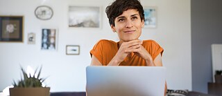 Eine Frau trägt eine orangefarbenes T-Shirt und sitzt gemütlich am Laptop. Sie denkt nach und lächelt.