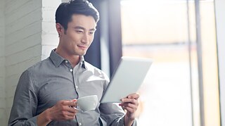 Person in grauem Hemd hält eine weiße Tasse in der linken und ein Tablet in der rechten Hand, sitzt vor einem Fenster mit hellem Licht. 