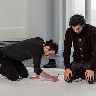 Zwei Männer knien in einem hellen Ausstellungsraum auf dem Boden und zeichnen konzentriert mit Stiften auf ein großes weißes Blatt Papier. 