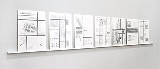 Das Foto zeigt eine weiße Wand, vor der eine schmale weiße Leiste angebracht ist. Auf der Leiste stehen sieben hochformatige, weiße Tafeln dicht nebeneinander. Jede Tafel ist mit grauen Zeichnungen, Textblöcken, Linien, Symbolen und kleinen grafischen Elementen versehen. Die Inhalte wirken fein, kleinteilig und variieren von Tafel zu Tafel. 