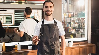 Ein Mann lächelt in die Kamera und hält eine Schere in der Hand. 
