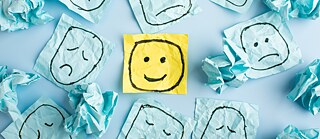 Ein gelber Smiley, umgeben von blauen, zerknüllten Haftnotizen mit traurigen Smileys darauf.