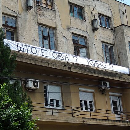 Ein altes, verwittertes Mehrparteienhaus mit bröckelnder Fassade und kaputten Fenstern. Über dem Balkon hängt ein langes weißes Banner mit der Aufschrift „ШТО Е ОВА? КИНО?“ in kyrillischen Buchstaben. Vor dem Gebäude verlaufen Strom- und Telefonleitungen, und im unteren Bereich sind Klimaanlagen angebracht.