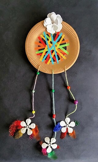 Ein selbstgebastelter Traumfänger aus Pappe mit Stofffäden, Holzkugeln und an den Fäden hängend bunte Federn sowie drei kleine runde Porträts von Sophie Scholl