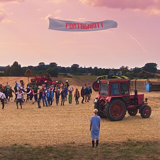 Szene aus dem Film „Rote Sterne überm Feld“: Menschen stehen auf einem Feld zwischen Traktoren; darüber fliegt ein Banner mit dem Wort ‚FORTSCHRITT‘