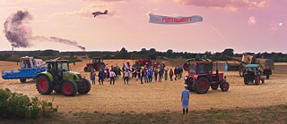 Szene aus dem Film „Rote Sterne überm Feld“: Menschen stehen auf einem Feld zwischen Traktoren; darüber fliegt ein Banner mit dem Wort ‚FORTSCHRITT‘