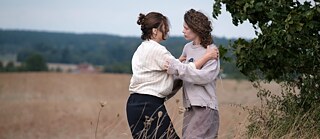 A dark and a light-haired woman stand in a field and hold each other's arms