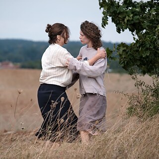 A dark and a light-haired woman stand in a field and hold each other's arms