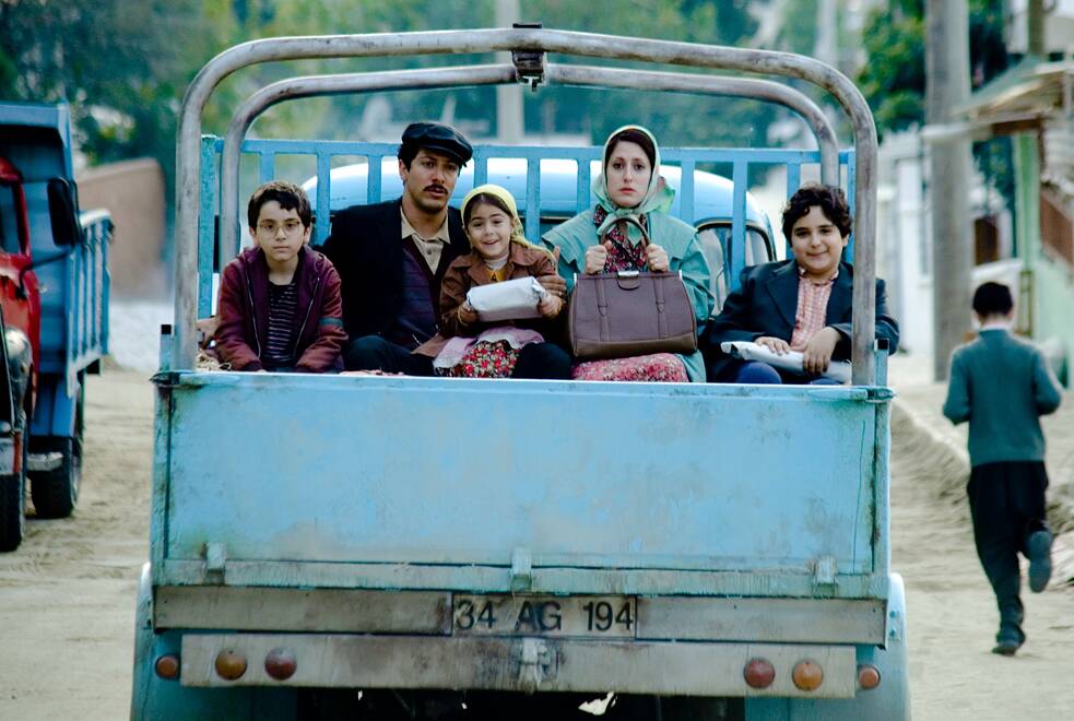 Sechs Personen sitzen auf der Ladefläche eines hellblauen Pick‑ups, der auf einer unbefestigten Straße steht. Drei Kinder und drei Erwachsene sitzen eng beieinander; zwei Kinder halten weiße Päckchen in den Händen. Ein Mann mit dunkler Jacke und Mütze sitzt in der Mitte, neben ihm eine Frau mit grünem Mantel und Kopftuch. Im Hintergrund sind weitere Fahrzeuge und eine Person zu sehen, die sich vom Bild wegbewegt.