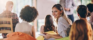 Frau sitzt mit anderen in Veranstaltung und lächelt in die Kamera