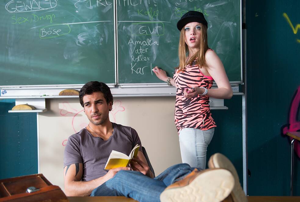 In einem Klassenzimmer steht eine junge Frau mit schwarz‑weiß gestreiftem Oberteil und schwarzer Kappe vor einer Tafel und erklärt etwas. Neben ihr sitzt ein junger Mann in grauem Shirt und Jeans lässig auf einem Tisch, die Füße hochgelegt, ein geöffnetes Buch in der Hand. Auf der Tafel stehen handgeschriebene deutsche Wörter und Begriffe. Die Szene wirkt lebhaft und zeigt einen informellen Unterrichtsmoment.