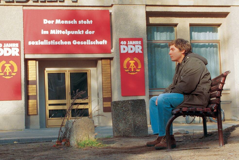 Ein Mann sitzt allein auf einer Bank vor einem Gebäude, das mit roten Bannern und Plakaten zum 40. Jubiläum der DDR geschmückt ist. Über dem Eingang hängen große Transparente mit sozialistischen Symbolen und Slogans. Der Mann trägt dunkle Kleidung und blickt nach vorn, während die Umgebung aus Betonflächen, Fenstern und etwas Vegetation besteht. Die Szene wirkt ruhig und leicht melancholisch.