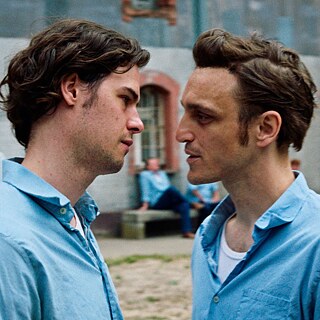 Kaks sinistes särkides meest vaatavad teineteisele silma. Nende taga istub mitu sama riietusega meest ühe hoone ees.