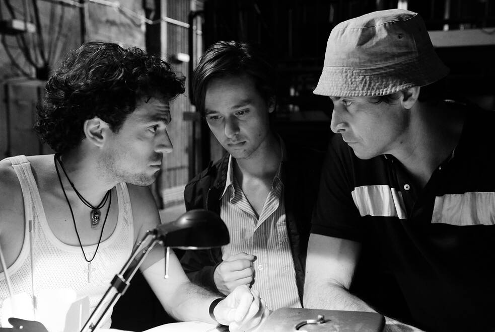 Drei Personen sitzen eng beisammen an einem Tisch, der von einer Schreibtischlampe beleuchtet wird. Die Szene ist in Schwarz‑Weiß gehalten. Links sitzt eine Person im Unterhemd mit mehreren Ketten, in der Mitte eine Person im Hemd, rechts eine Person mit Fischerhut und Poloshirt. Alle drei beugen sich konzentriert über den Tisch, während im Hintergrund Rohre und Metallstrukturen zu sehen sind. Die Stimmung wirkt ernst und nachdenklich.