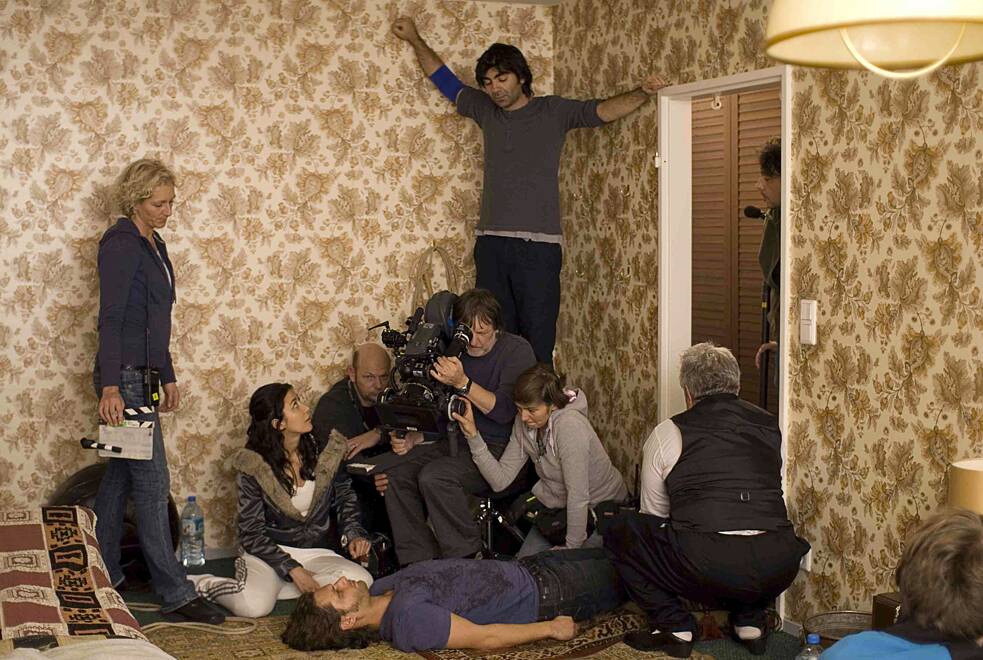 Mehrere Personen arbeiten in einem kleinen, mit gemusterter Tapete versehenen Raum an einer Filmszene. Eine Kamera ist auf zwei Schauspieler gerichtet, von denen einer am Boden liegt und der andere daneben sitzt. Crewmitglieder halten Ausrüstung, richten Licht und Ton aus und stehen dicht um die Szene herum. Die Atmosphäre wirkt konzentriert und geschäftig, typisch für eine Filmproduktion in engem Raum.