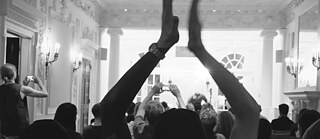 Black and white photo of a person in silohuette clapping with their hands over their head in the Boston Institut Grand Hall