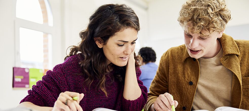 Ein Mann und eine Frau befinden sich in einem Klassenraum. Sie sind in ein Gespräch vertieft. Die Frau stützt sich mit dem Ellenbogen auf den Tisch vor ihnen, während der Mann ihr mit seinem Kugelschreiber etwas auf einem Arbeitsblatt zeigt.