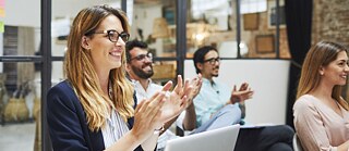 A group of people seated in a modern office space clapping during a presentation or meeting, with a laptop open in the foreground and large glass partitions in the background