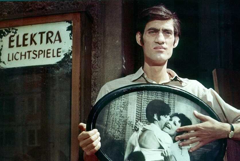 Ein Mann steht vor einem Gebäude mit dem Schriftzug „Elektra Lichtspiele“ und hält einen großen ovalen Rahmen mit einem schwarz‑weißen Foto eines sich umarmenden Paares. Er trägt ein helles Hemd und eine Brille. Hinter ihm sind Türen und Fenster des Gebäudes zu sehen. Der Rahmen mit dem Foto dominiert das Bild und lenkt den Blick auf das dargestellte Paar.