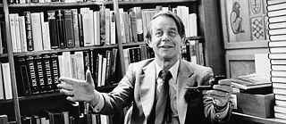 Siegfried Lenz, sitting in front of a bookshelf with a friendly smile, holding a pipe in one hand and extending his open arms toward the viewer.