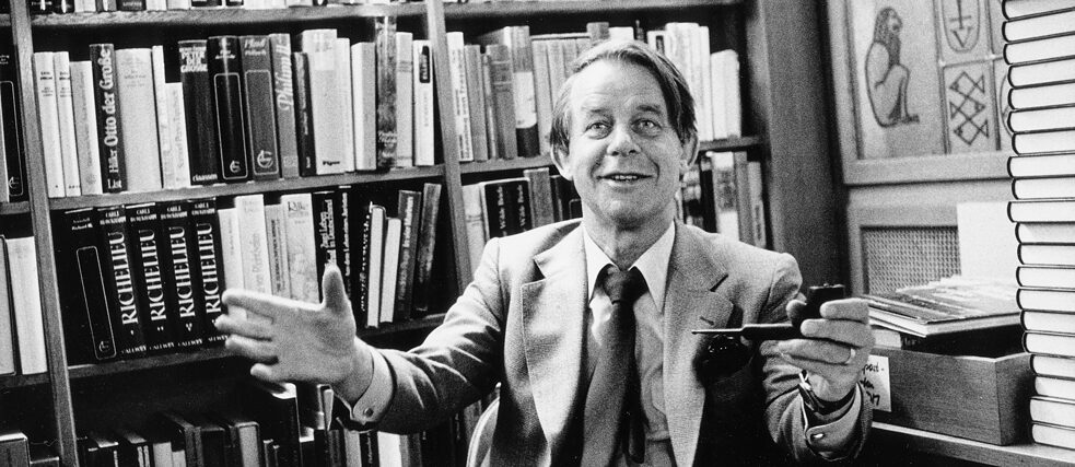 Siegfried Lenz, sitting in front of a bookshelf with a friendly smile, holding a pipe in one hand and extending his open arms toward the viewer.