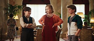 Drei Kinder stehen gemeinsam in einem eleganten Raum mit hohen Marmorsäulen, warmem Licht und klassischem Mobiliar.