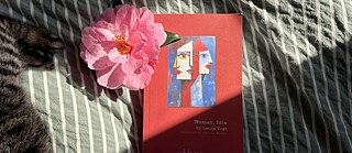 The book 'Woman, Idle' lies on a striped bedsheet alongside a pink camellia blossom