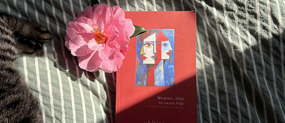 The book 'Woman, Idle' lies on a striped bedsheet alongside a pink camellia blossom
