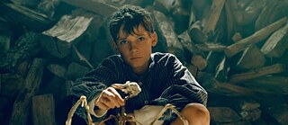 Szene aus der Literaturverfilmung „Deutschstunde“ von Siegfried Lenz.  Es zeigt die Hauptfigur Siggi Jepsen als Kind, dargestellt von Levi Eisenblätter, vor einem Stapel Brennholz. 