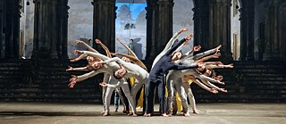 Tänzer*innen in synchroner Gruppenpose auf einer Bühne vor historischen Arkaden.