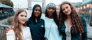 Vier junge Menschen stehen eng beieinander an der Themse in London, mit der Tower Bridge im Hintergrund. Sie tragen Freizeitkleidung und posieren vor einer städtischen Kulisse am Wasser.