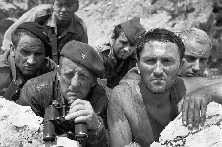 Sechs Männer in militärischer Kleidung ducken sich hinter Felsen in einer rauen Außenlandschaft. Einige tragen Mützen mit Sternsymbolen, andere einfache Uniformkappen. Ein Mann im Vordergrund hält ein Fernglas, ein anderer eine Pistole; alle blicken konzentriert in dieselbe Richtung. Die Szene wirkt angespannt, mit dicht gedrängten Körperhaltungen und unruhigem Gelände im Hintergrund.