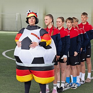 Lehrerin im Fußballkostüm steht vor einer Reihe von Jugendlichen in Sportkleidung auf dem Fussballfeld.