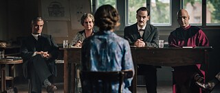 Eine Frau sitzt mit dem Rücken zur Kamera und blickt auf eine Gruppe von vier Personen, die hinter einem Holztisch in einem schwach beleuchteten Raum sitzen.