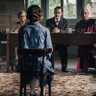  Eine Frau sitzt mit dem Rücken zur Kamera und blickt auf eine Gruppe von vier Personen, die hinter einem Holztisch in einem schwach beleuchteten Raum sitzen.