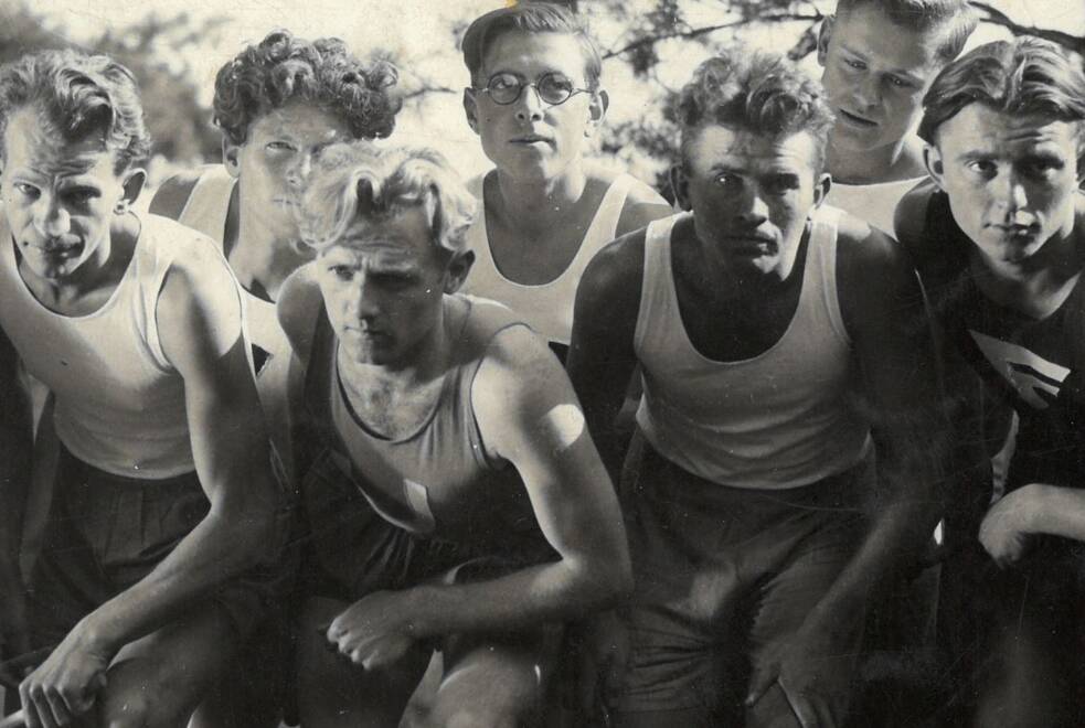 Mehrere junge Männer in Sportkleidung hocken in Startposition auf einer Laufbahn. Sie tragen ärmellose Shirts und kurze Hosen. Die Aufnahme ist in Schwarz‑Weiß gehalten und zeigt konzentrierte, entschlossene Gesichter. Die Szene wirkt wie der Beginn eines Wettlaufs und vermittelt Energie, Anspannung und den sportlichen Geist der Zeit.